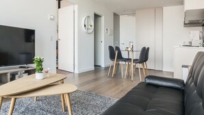 A modern living room with a black leather sofa, a wooden coffee table, and a TV. There is a dining area with a table and chairs, and a kitchen in the background.
