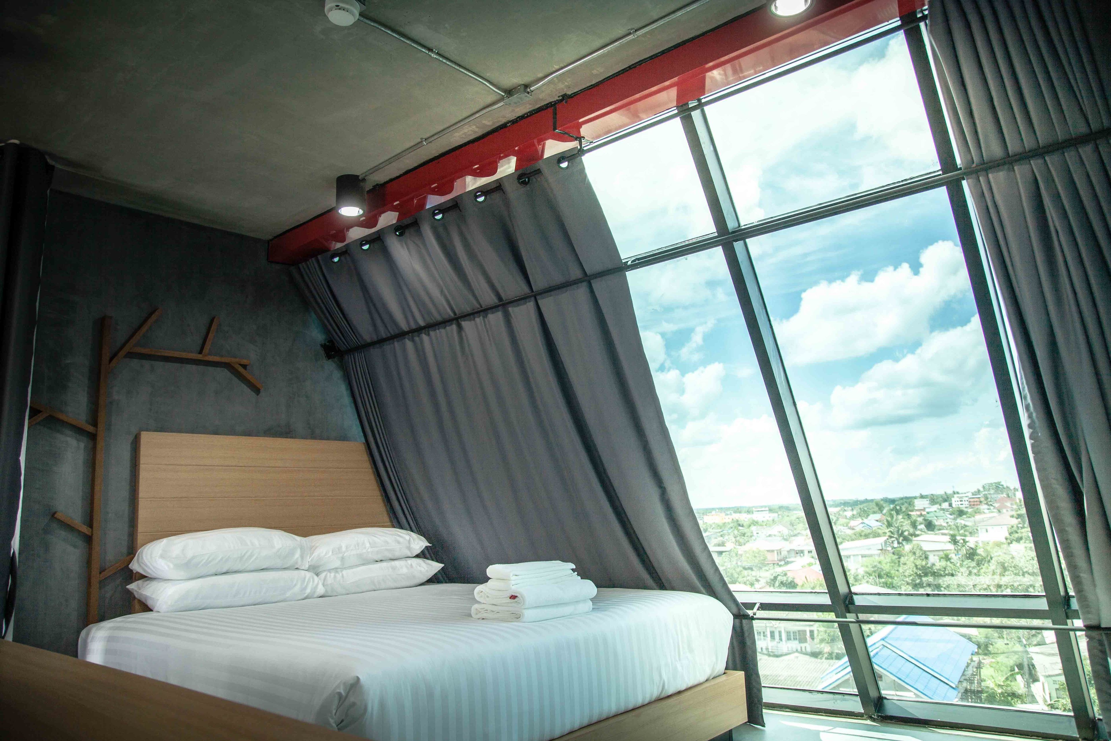 A modern hotel room with a large bed, a view of the city through large windows, and a red accent on the ceiling.