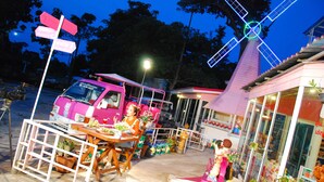 Uma carrinha rosa estacionada junto a um quiosque à beira da estrada, com uma mesa e cadeiras, rodeado por vasos de plantas e uma decoração de moinho de vento.