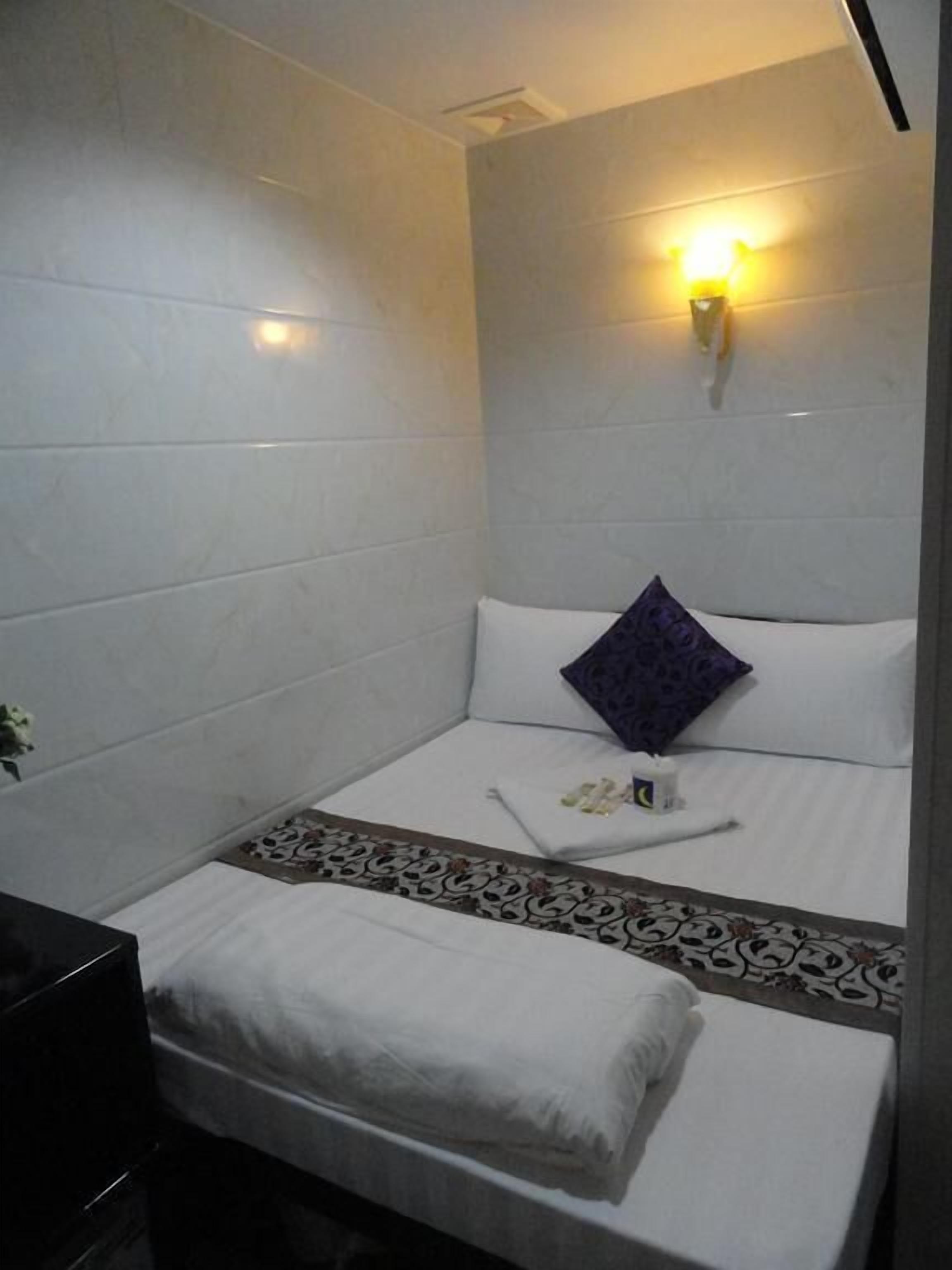 A small, neatly arranged room with a bed, a bedside table, and a wall-mounted light fixture.