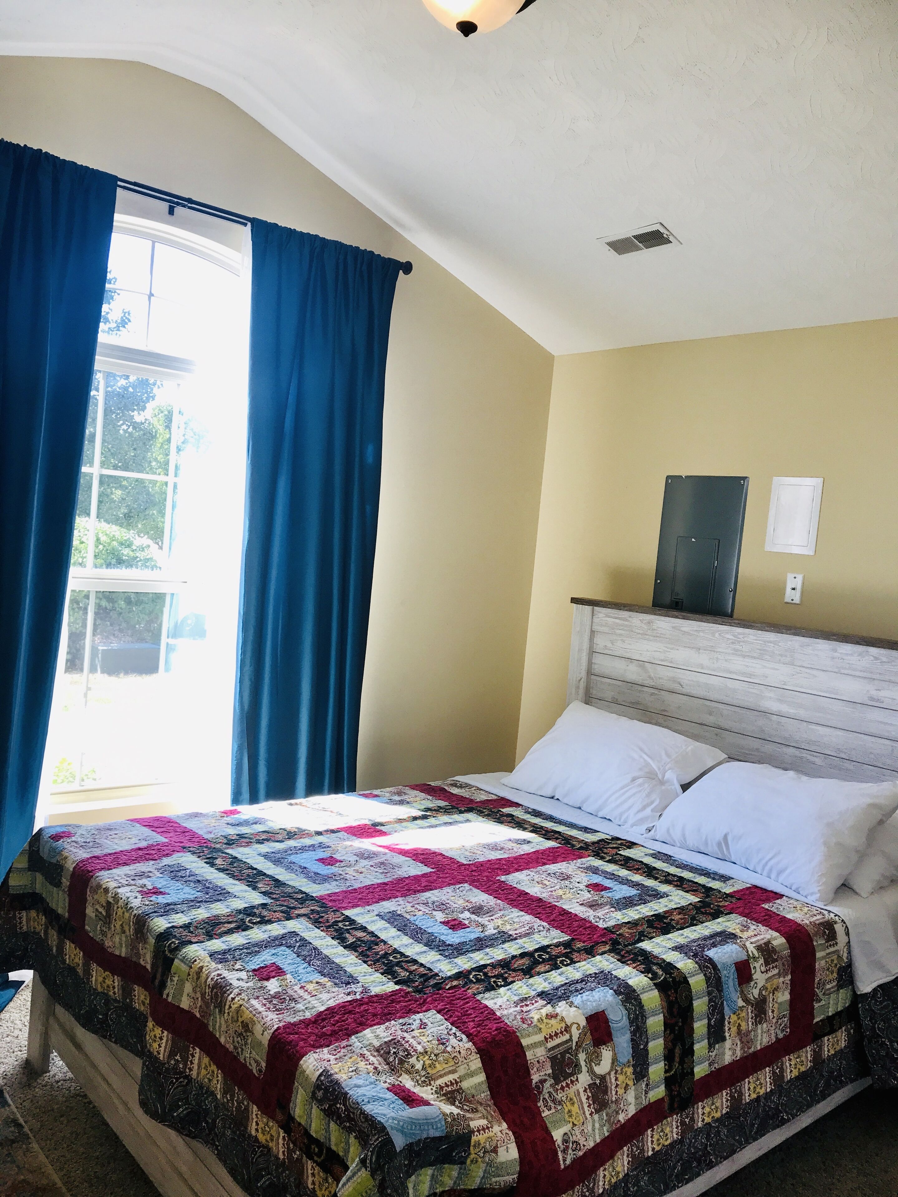 Un dormitorio con una cama grande, una ventana con cortinas azules, un cabecero de madera y una lámpara fijada a la pared.