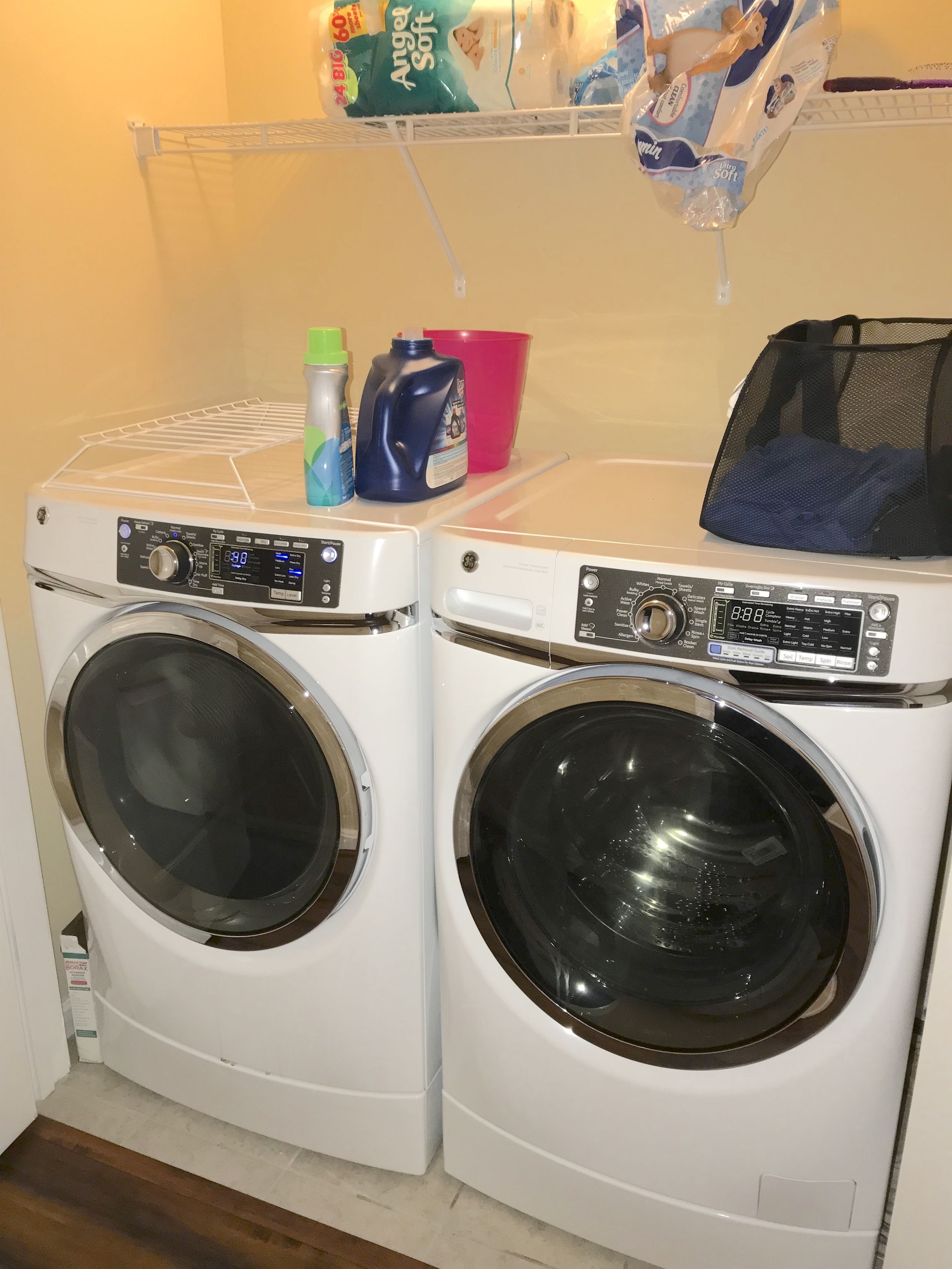 Two front-loading washing machines with control panels, a drying rack, and laundry supplies on a shelf.