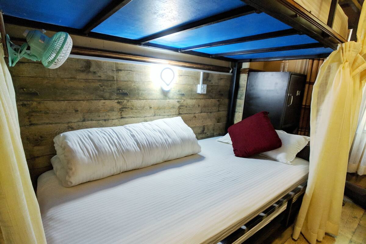 A bunk bed with a white mattress, a red pillow, and a white blanket. There is a ceiling fan above the bed, and a wooden wall behind it.