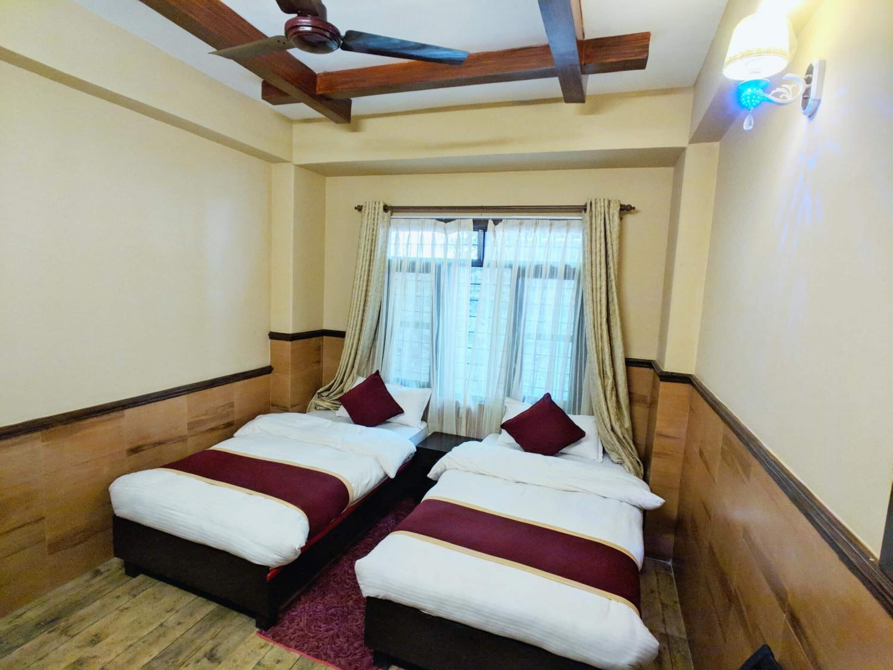 Ein Hotelzimmer mit zwei Betten, einem Deckenventilator, einem Fenster mit Vorhängen und einer wandmontierten Lampe.