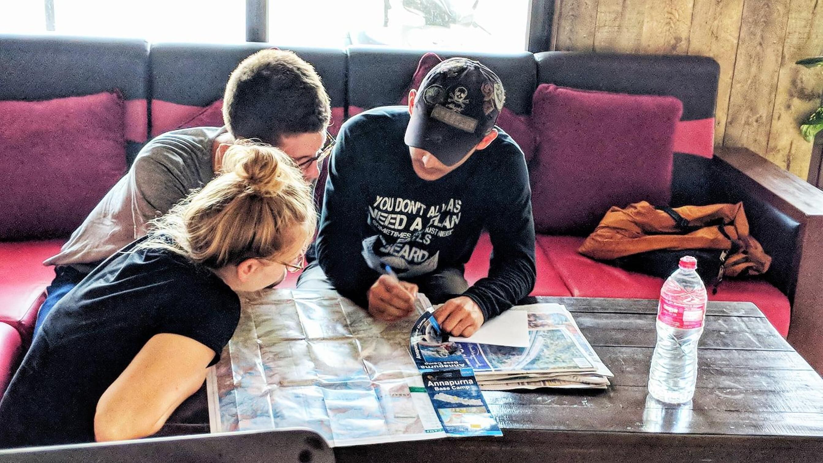 Three people sitting on a red couch, one person writing on a map, and a bottle of water on a wooden table.