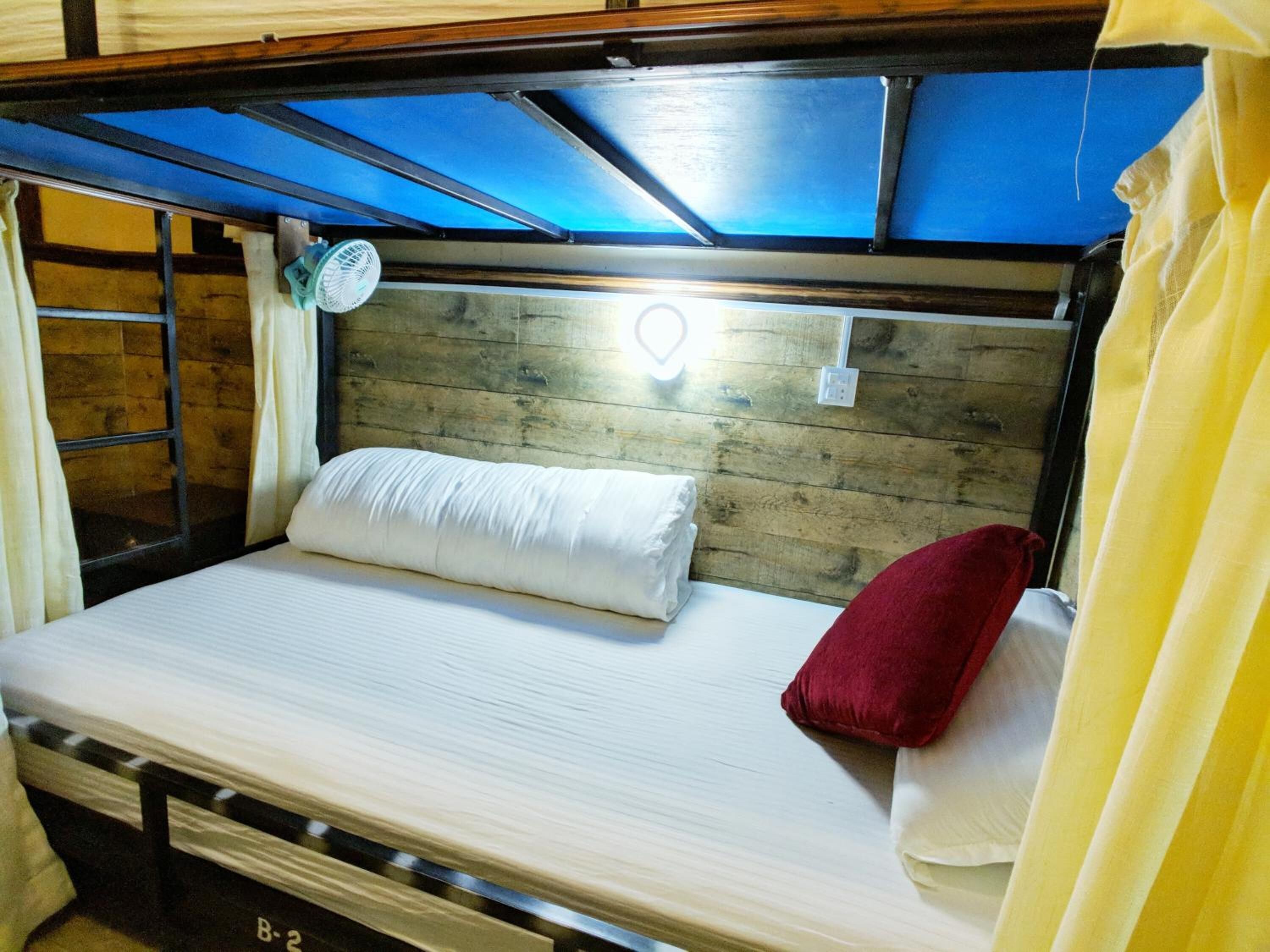 Una cama litera con cabecera de madera, un colchón blanco, una almohada roja y un pequeño ventilador.