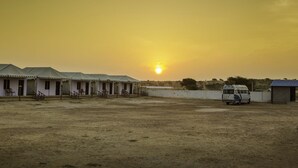 A row of single-story buildings with a dirt ground, a parked van, and a clear sky with the sun setting.