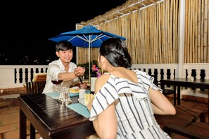 A couple dining at an outdoor table with a blue umbrella, surrounded by bamboo walls and a white balustrade. - Hotel De L'Homme (Phnom Penh)