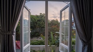 An open window with curtains, revealing a view of a garden with various plants and a brick wall. - Hotel De L'Homme (Phnom Penh)