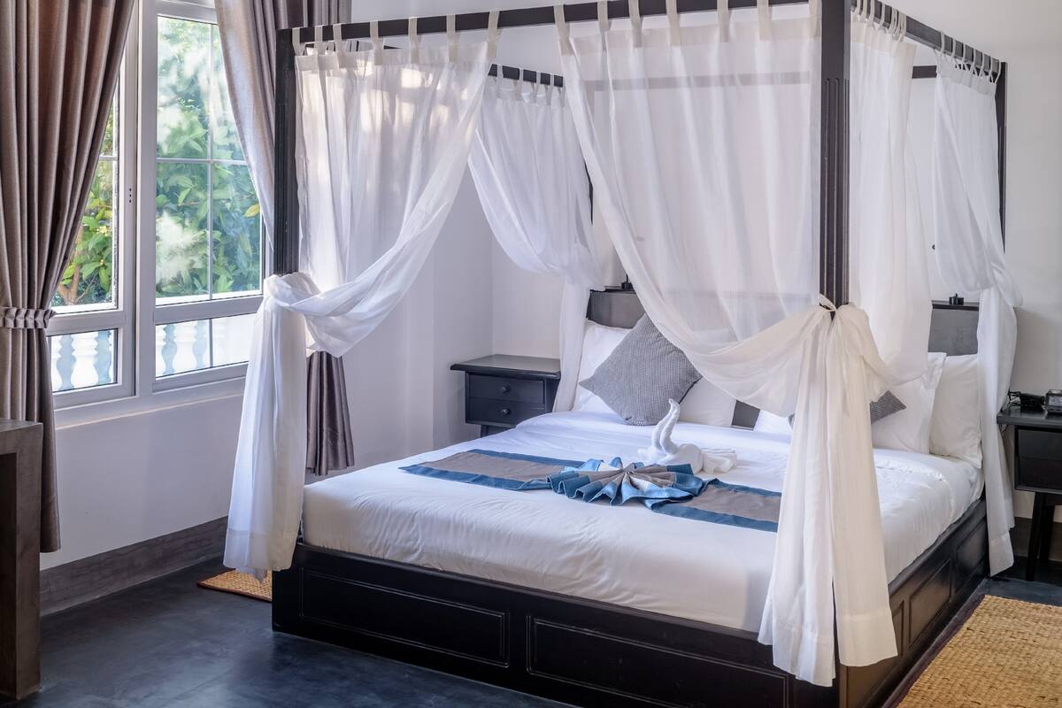 A four-poster bed with white bedding and a blue accent, a bedside table with a lamp, and a view of greenery through a window.