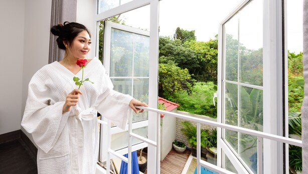 A person in a white robe holding a rose, standing by a large window with a view of a garden and a pool. - Hotel De L'Homme (Phnom Penh)