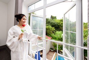 Uma pessoa vestida com um robe branco segura uma rosa, em frente a uma grande janela com vista para um jardim e uma piscina.