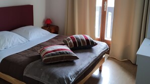 Quarto com cama, almofadas, mesa de cabeceira, abajur e janela com cortinas.