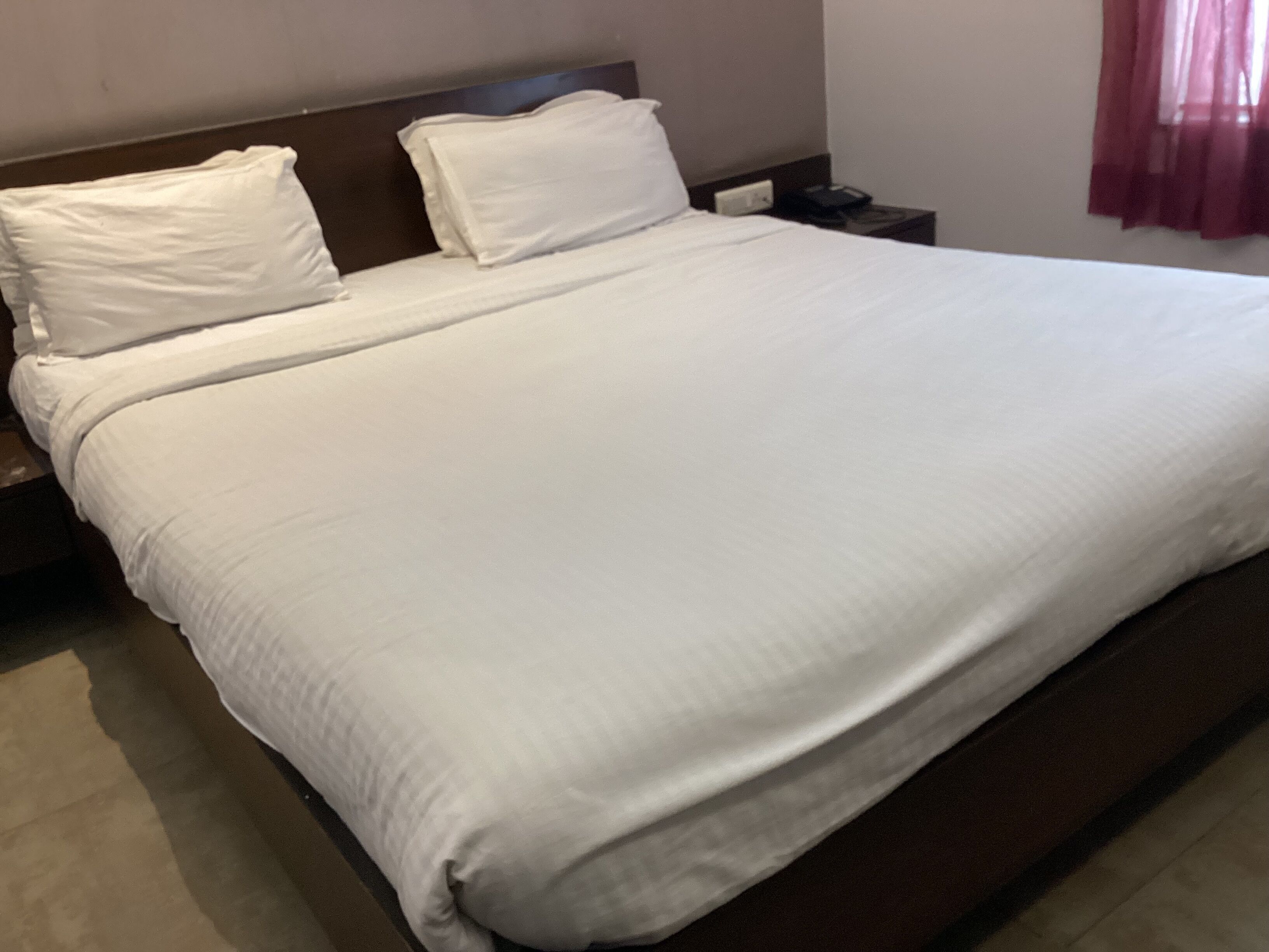 A hotel room with a large bed, a wooden headboard, a nightstand, and a red curtain.
