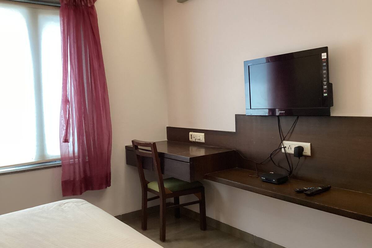 Una habitación de hotel con una cama, un escritorio con una silla, un televisor montado en la pared y una ventana con cortina.