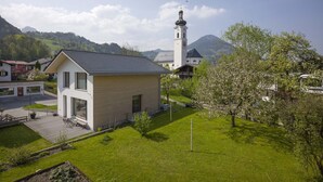 Ein modernes Gebäude mit geneigtem Dach, eine kleine Gartenanlage mit einer Holzterrasse und im Hintergrund eine Kirche mit einem Glockenturm.