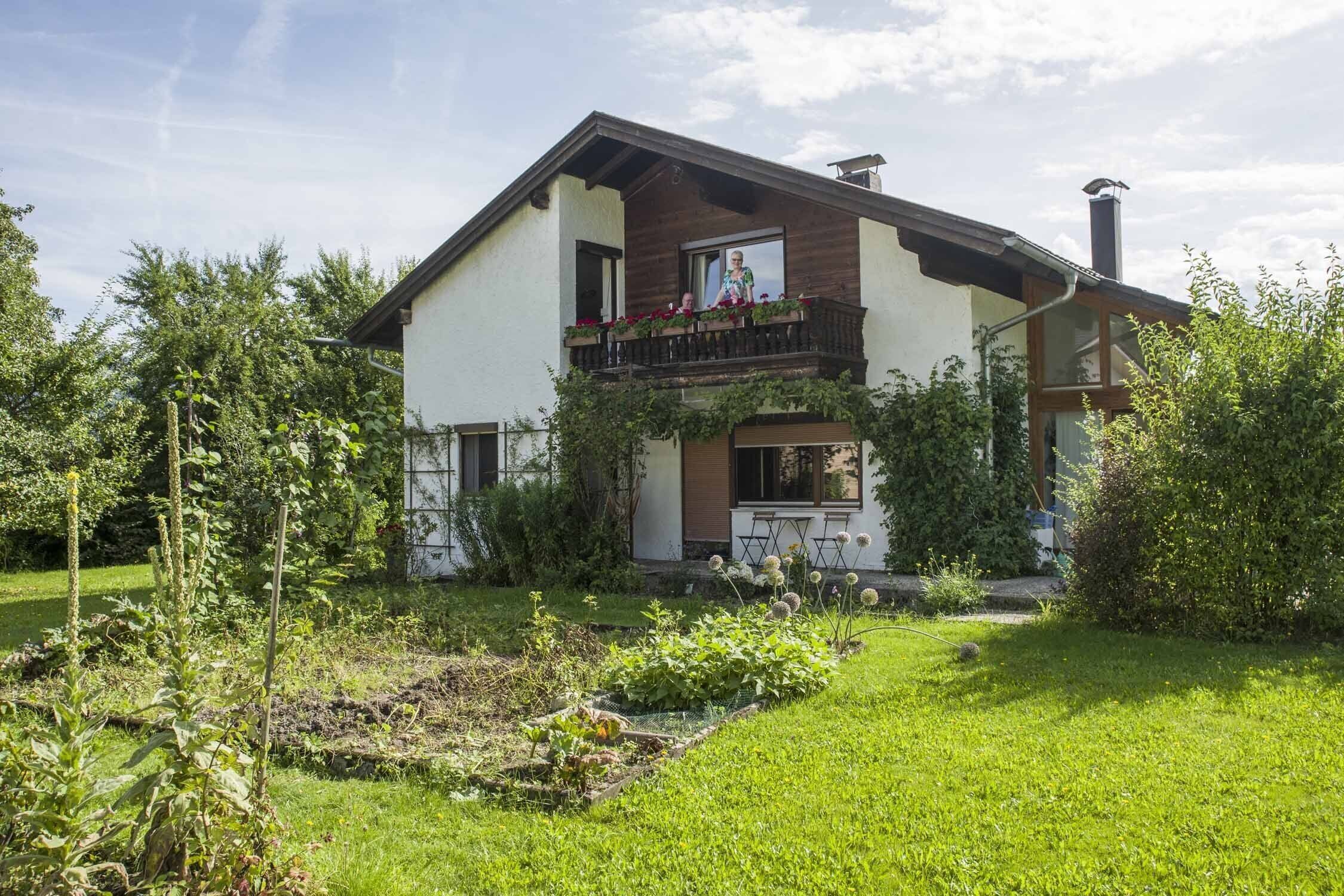 Una casa de dos pisos con un balcón, rodeada de un jardín cuidado y árboles.
