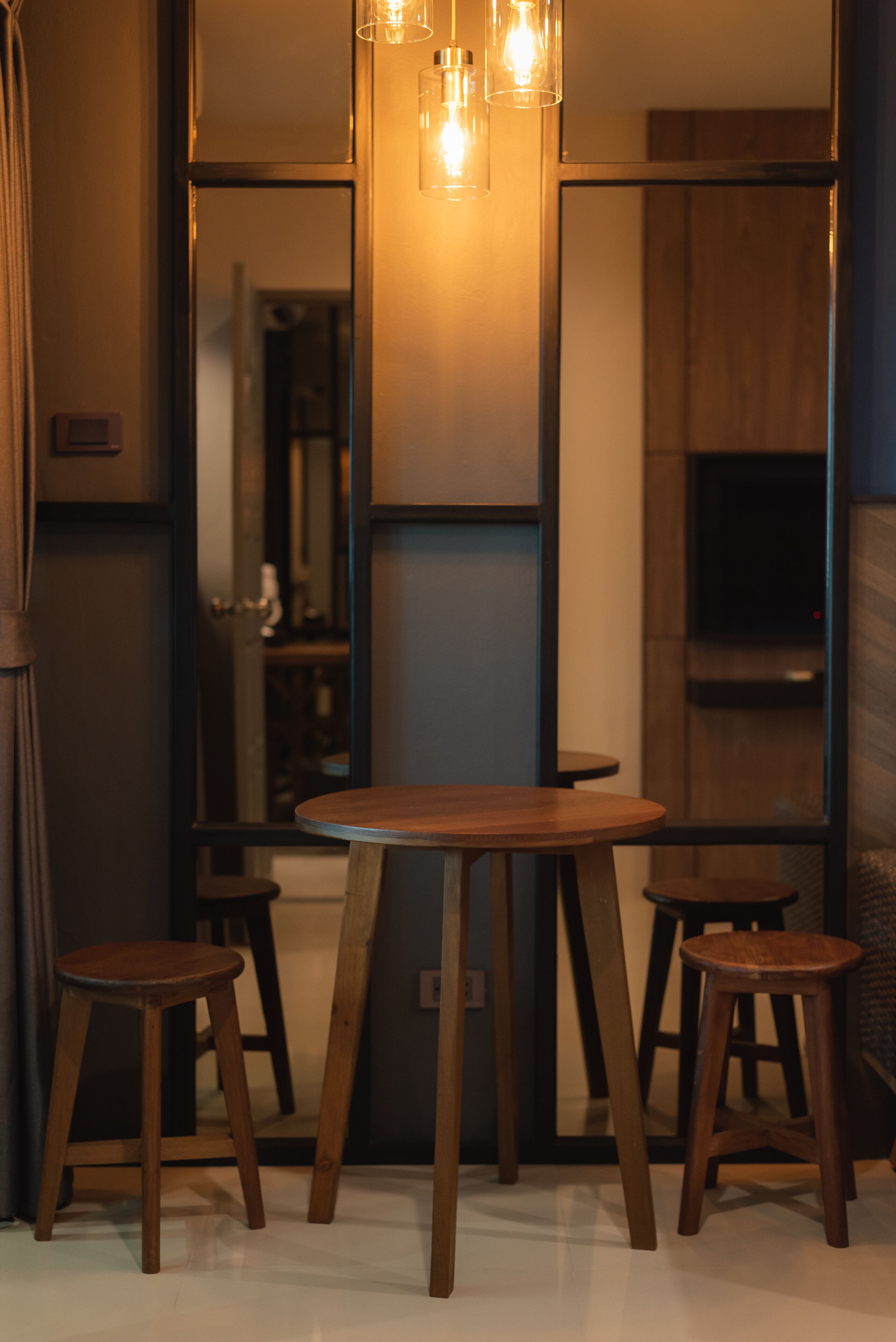 A modern interior with a round wooden table and stools, a hanging light fixture, and a large mirror reflecting the room.