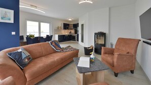 Un salon moderne avec un canapé marron, une table basse et un fauteuil marron. On aperçoit une cuisine à l’arrière-plan, avec des placards et une cuisinière.