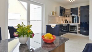 Une cuisine moderne avec des meubles de couleur sombre, un îlot central en verre et un coin repas agrémenté d’un vase de fleurs et d’un panier de fruits.
