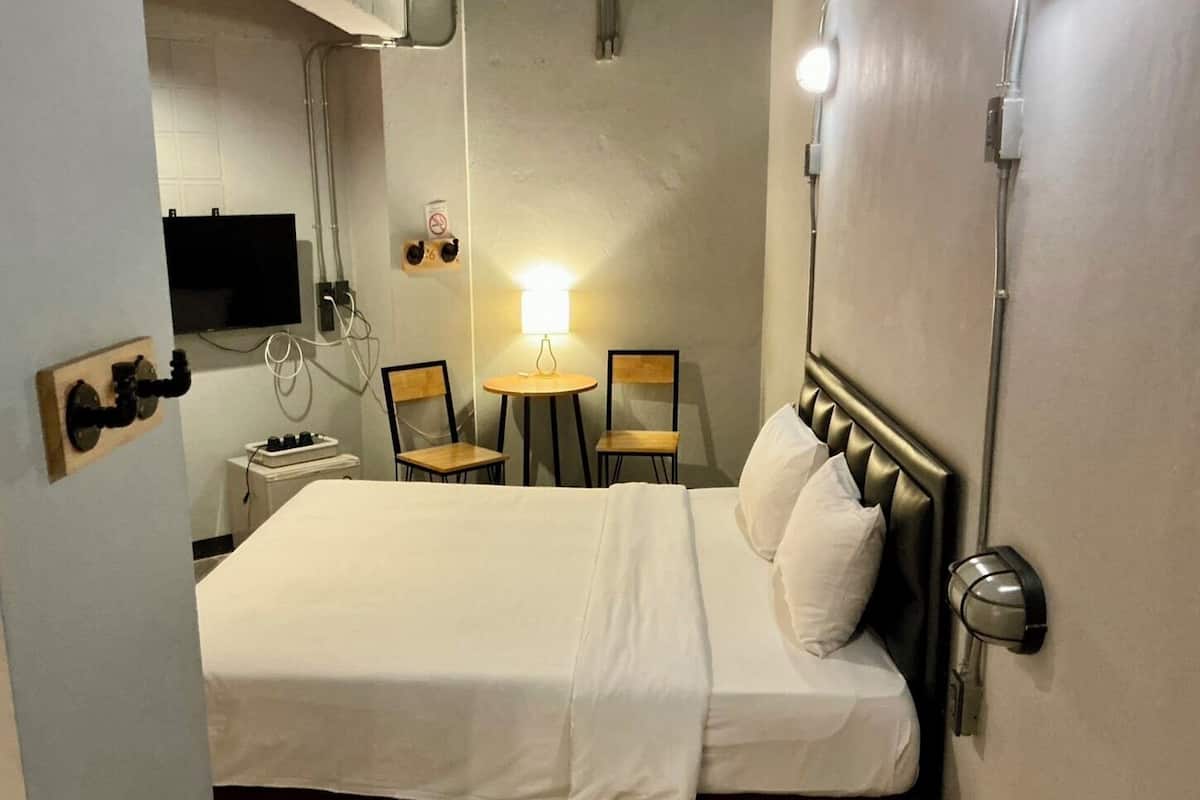A hotel room with a bed, a desk, a chair, a small table, and a TV mounted on the wall.