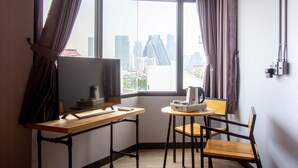 A room with a desk, chair, and a small table with a kettle and cups. A large window offers a view of a cityscape. - Rest24 Residence (Bangkok)