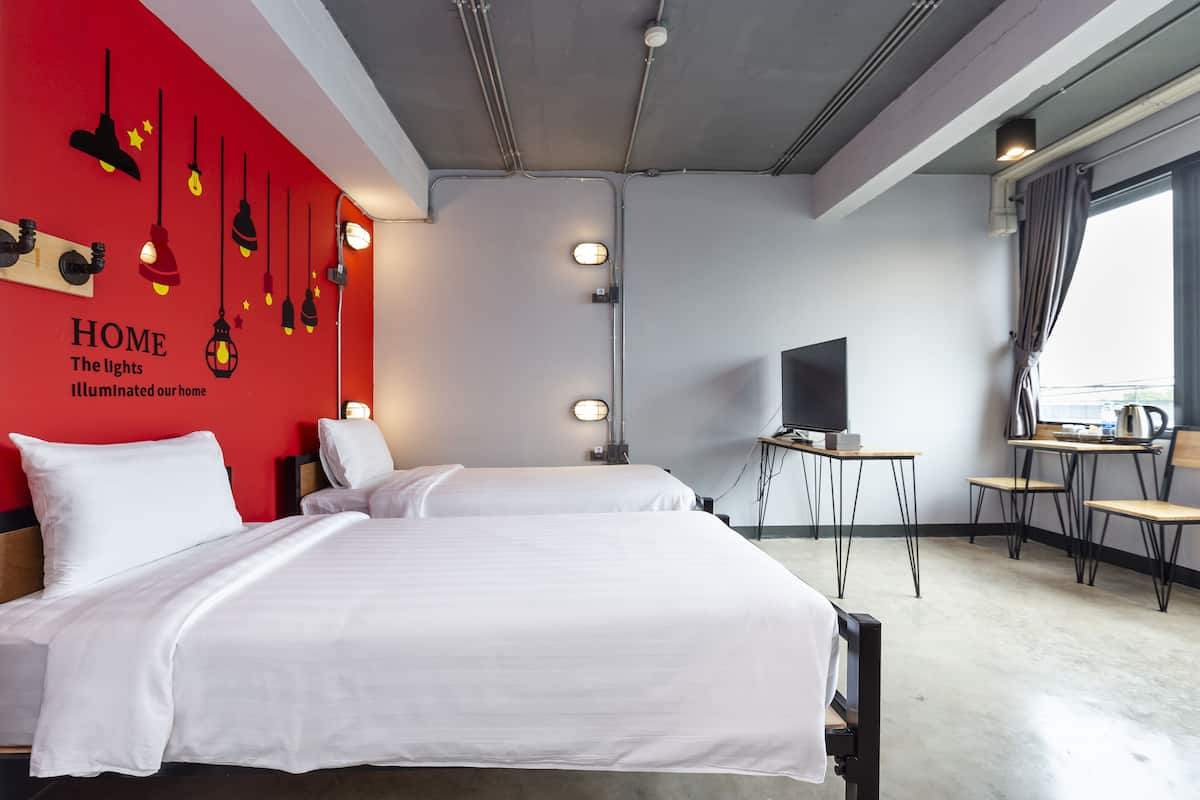 A hotel room with two beds, a TV, a small table with a kettle, and a red wall with hanging lights.