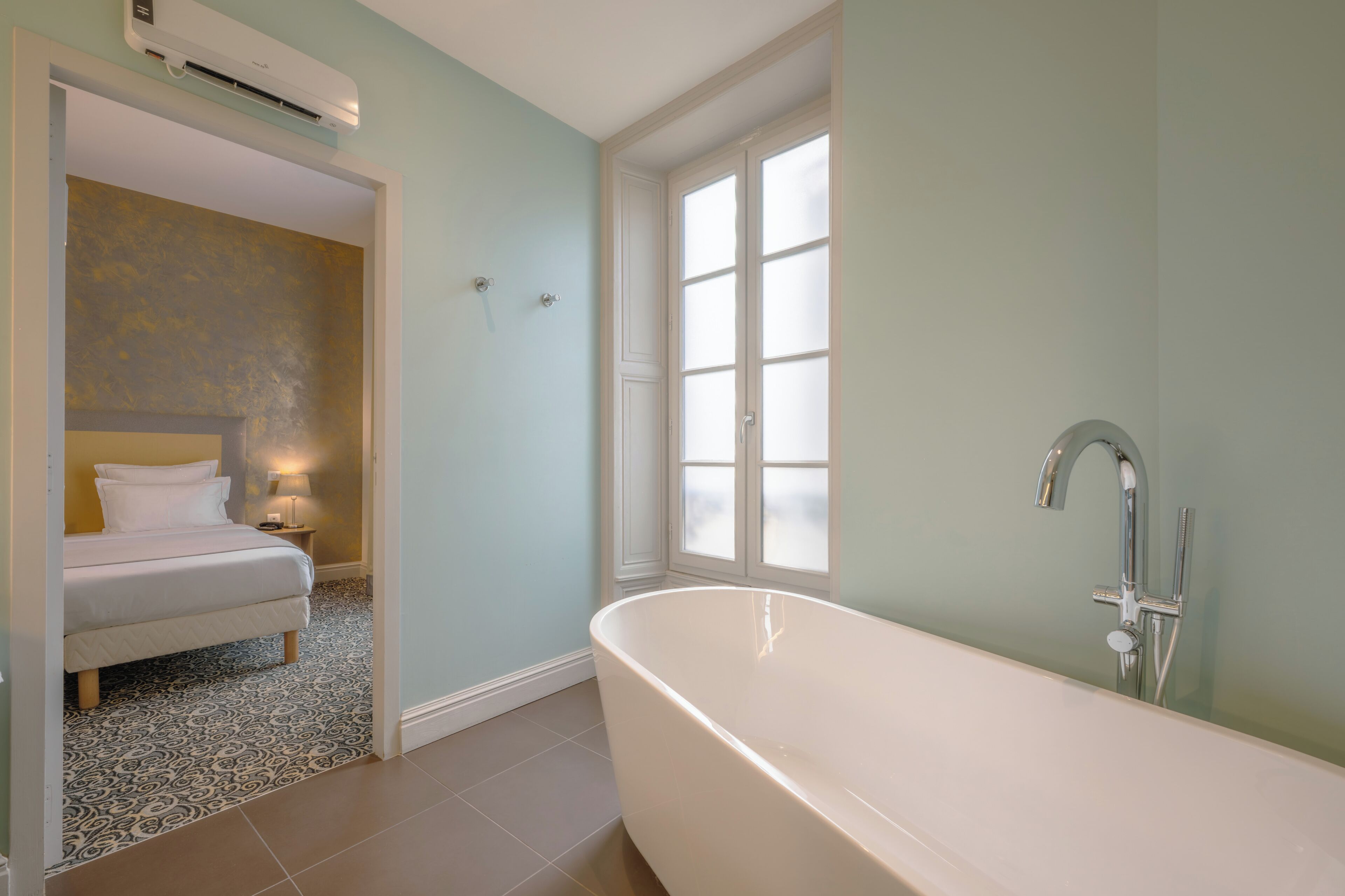 A bathroom with a bathtub, a window, and a bed with a nightstand.