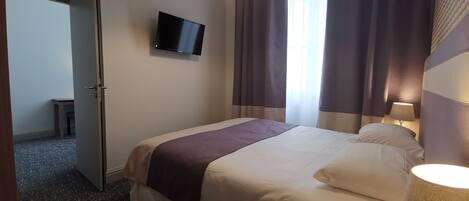 Habitación de hotel con cama, televisión instalada en la pared, lámpara y ventana con cortinas.
