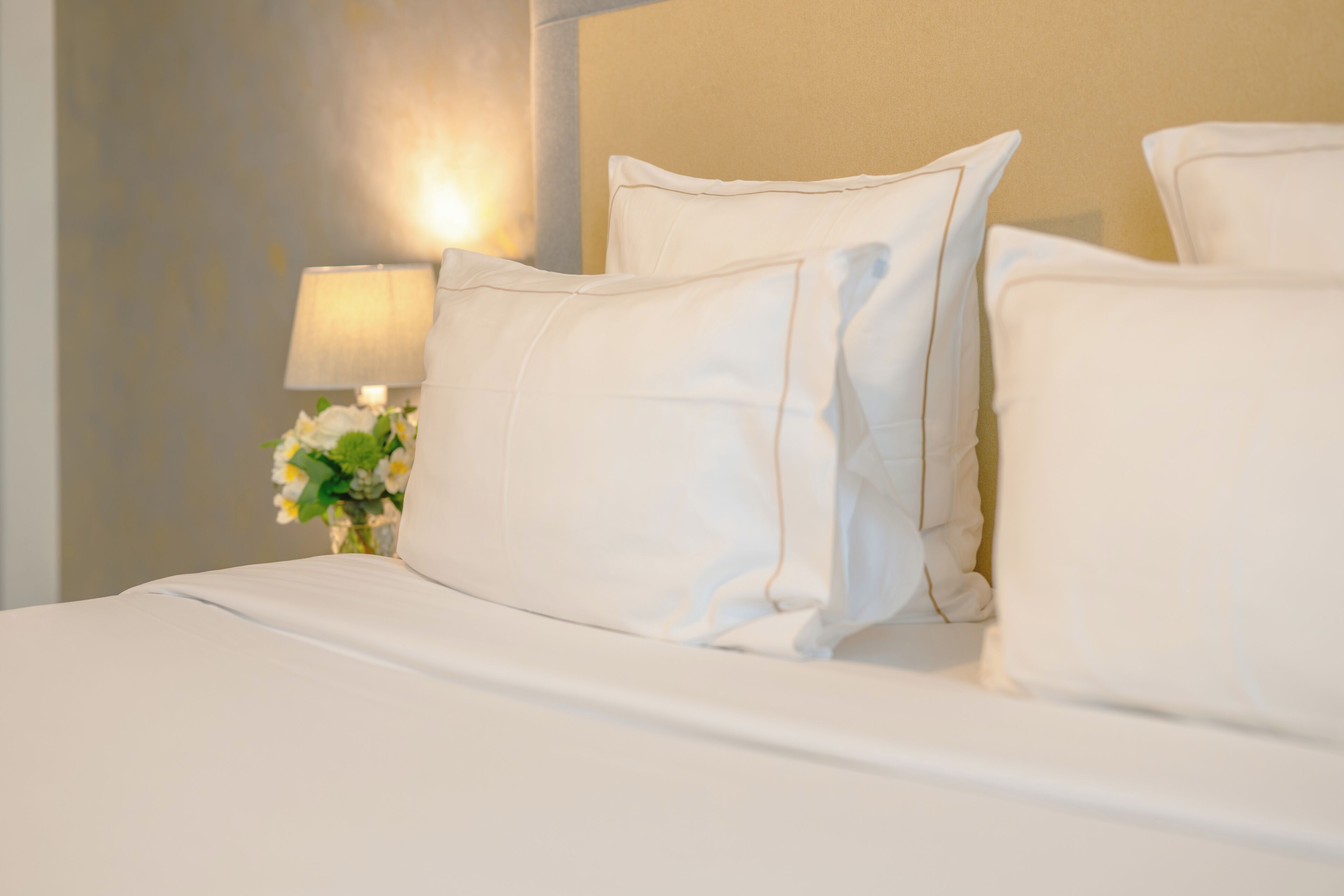 A neatly made bed with white linens and pillows, a bedside lamp, and a vase with flowers.