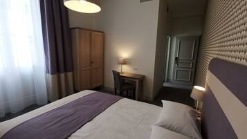 A hotel room with a bed, a desk, a chair, a wardrobe, and a door leading to another room.