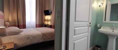 Habitación de hotel con cama, lámpara de noche, escritorio, silla y baño con lavamanos y espejo.