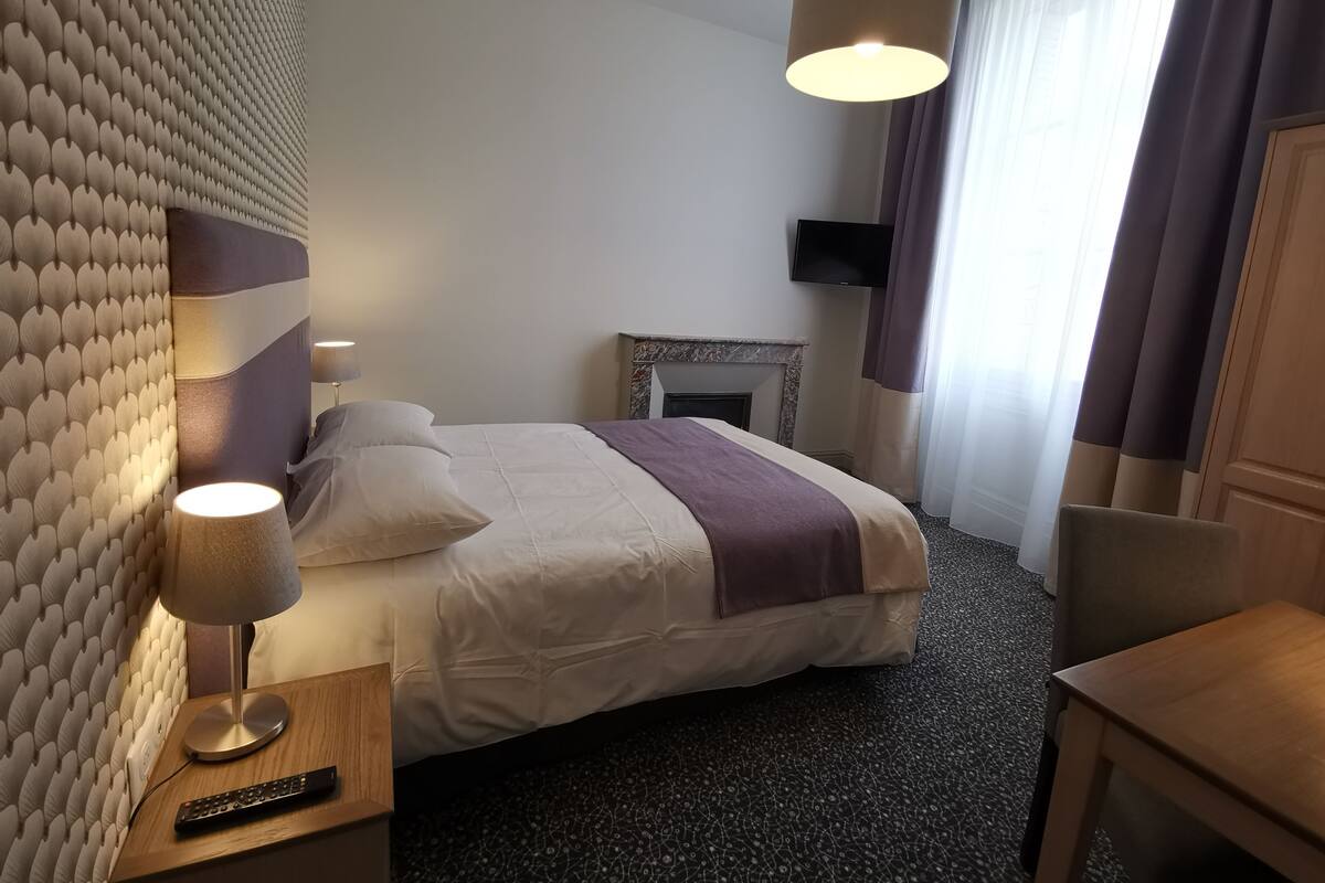 Una habitación de hotel con cama, lámpara de noche, control remoto, chimenea y televisión en la pared.