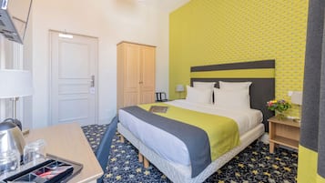 A hotel room with a bed, a desk, a chair, a wardrobe, and a nightstand with a lamp and flowers.