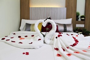 Una habitación de hotel con una cama bien hecha, una disposición de toallas en forma de corazón y pétalos de rosa esparcidos.