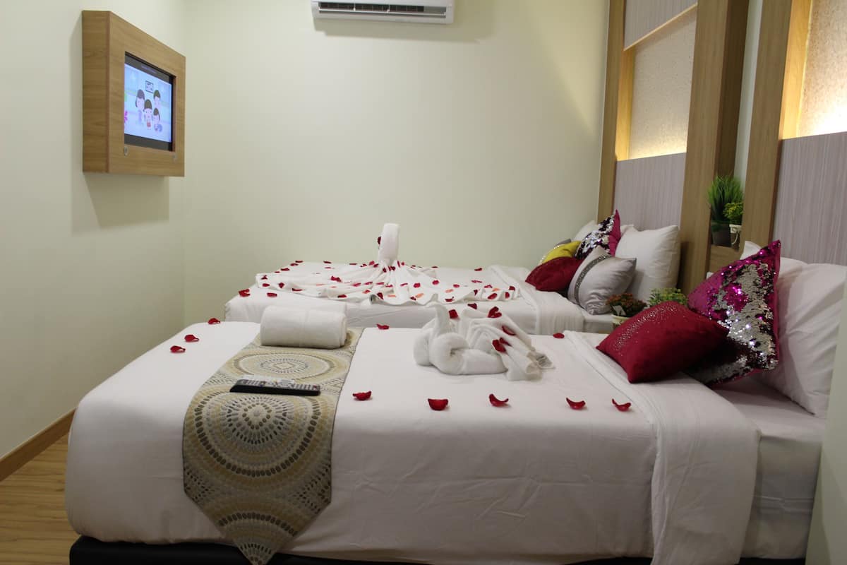 A hotel room with two beds, each adorned with white linens, rose petals, and decorative swans. A television is mounted on the wall, and a small plant is visible on the bedside table.