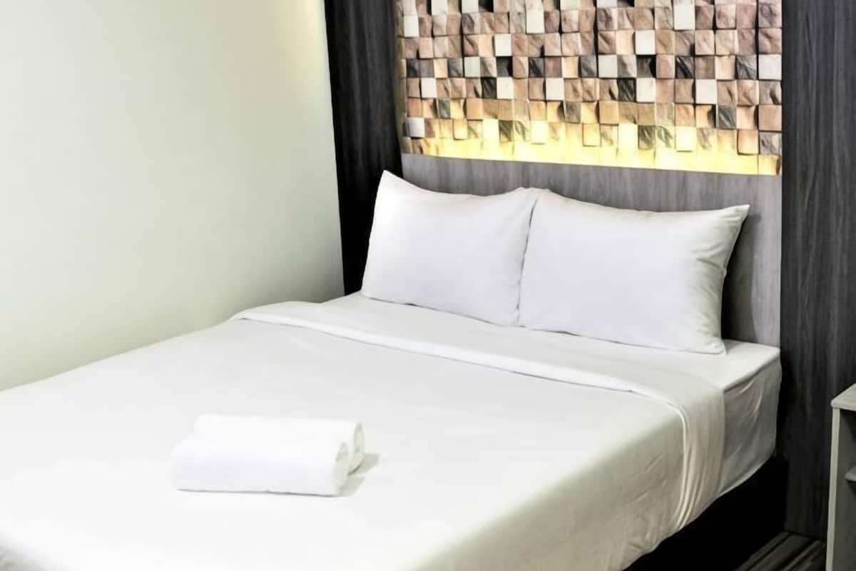 A neatly made bed with white linens and pillows, a folded white towel, and a headboard with a geometric pattern.
