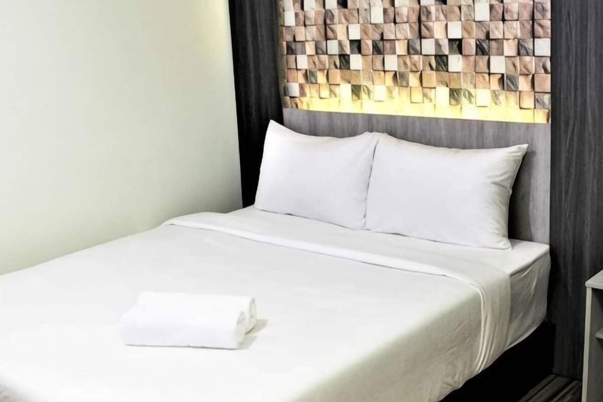 A neatly made bed with white linens and pillows, a folded white towel, and a headboard with a geometric pattern.