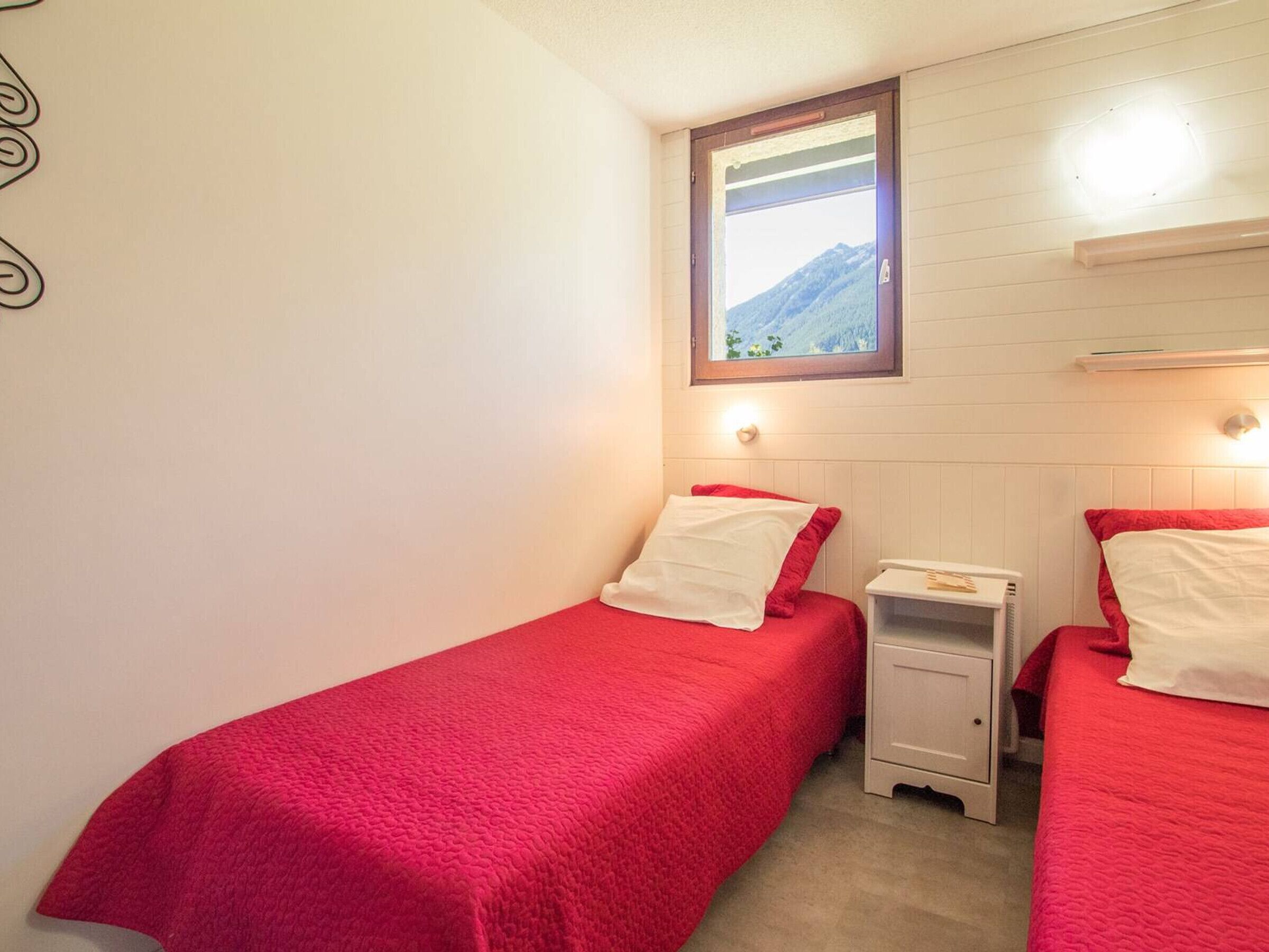 A small, cozy room with two single beds, each with a red bedspread and white pillows. There is a small bedside table with a lamp, a window with a view of a mountain, and a wall-mounted shelf above the beds.