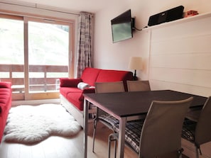 Una habitación con un sofá rojo, una mesa de comedor con sillas, un televisor montado en la pared y vistas a través de una puerta corrediza.
