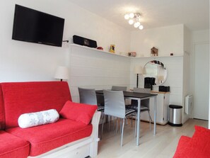 Ein modernes Wohnzimmer mit einem roten Sofa, einem Esstisch mit Stühlen, einem an der Wand befestigten Flachbildfernseher und einer Deckenleuchte.