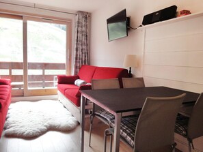Um quarto com um sofá vermelho, uma mesa de jantar com cadeiras, uma televisão fixada na parede e vista através de uma porta deslizante.