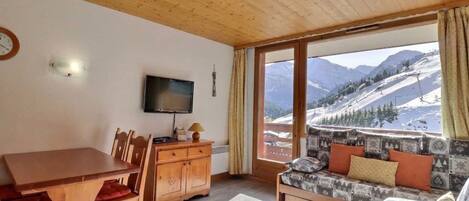 A cozy room with a wooden ceiling, a dining table with chairs, a sofa with patterned cushions, a flat-screen TV, and a view of snow-covered mountains.