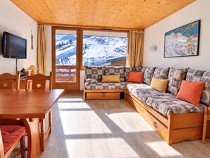 A cozy room with a wooden ceiling, a sofa with patterned cushions, a dining table with chairs, and a television.