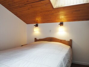 Une chambre avec un plafond en bois, deux lampes fixées au mur et un lit recouvert d’une couette blanche.