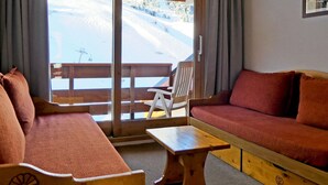 Ein Zimmer mit einer hölzernen Bank, einem Sofa und einem Tisch. Eine Glastür führt zu einem Außenbereich mit einer Holzterrasse und einem Stuhl.