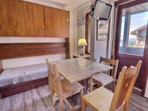 A compact room with a wooden headboard bed, a wooden dining table with chairs, a TV mounted on the wall, and a window with a view of buildings. - 1* Méribel Ski-in/Ski-out Studio - WiFi (Méribel)