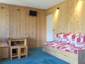 A cozy room with a wooden wall, a bed with a red and white quilt, a small wooden table, and a TV mounted on the wall.