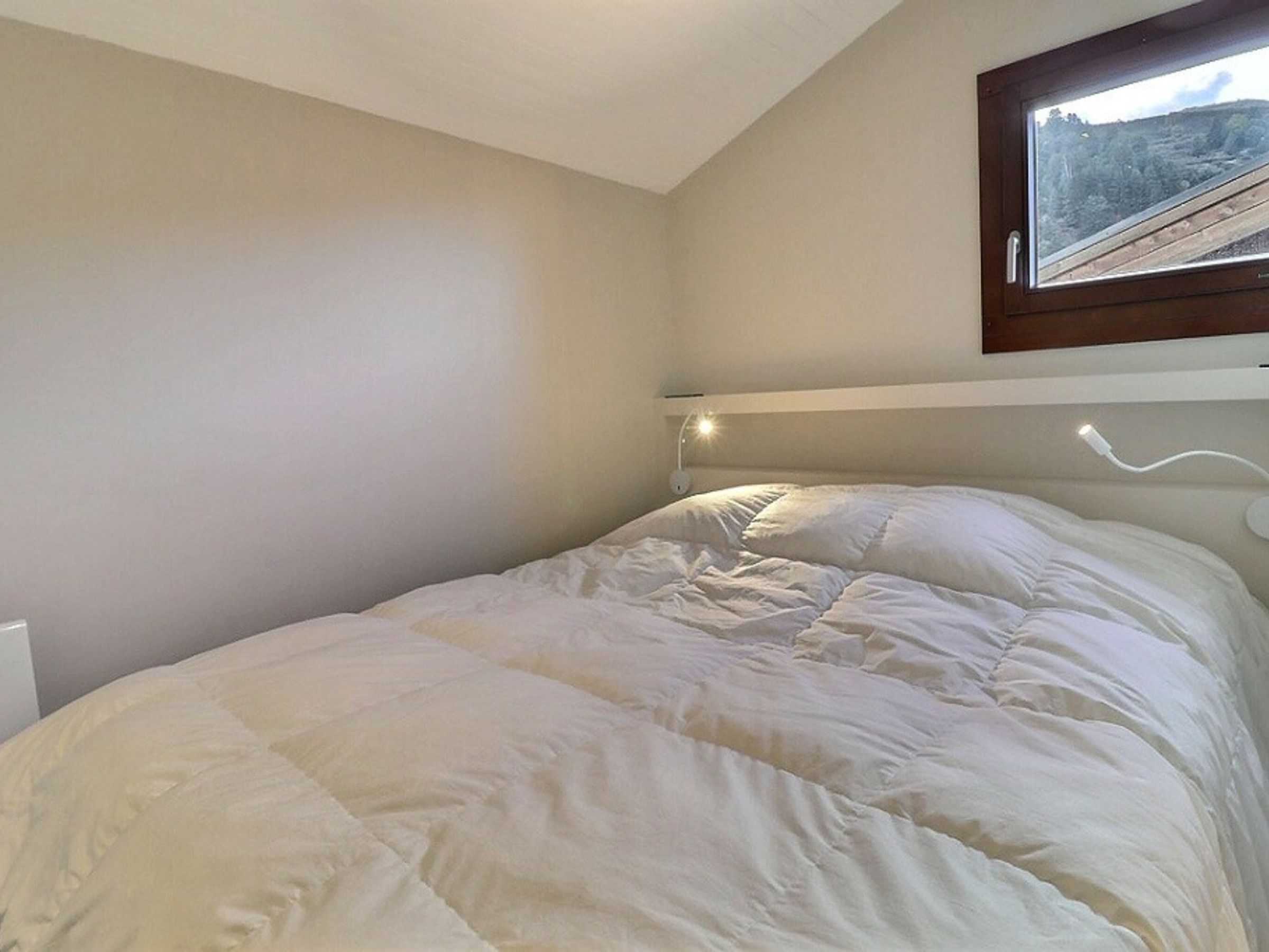 Un lit simple avec une couette blanche, dans une pièce dotée d’un plafond en pente et d’une fenêtre donnant sur un paysage boisé.