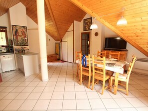 Uma sala de jantar espaçosa com cadeiras e mesa de madeira, uma cozinha com frigorífico e micro-ondas, e uma televisão ao fundo.